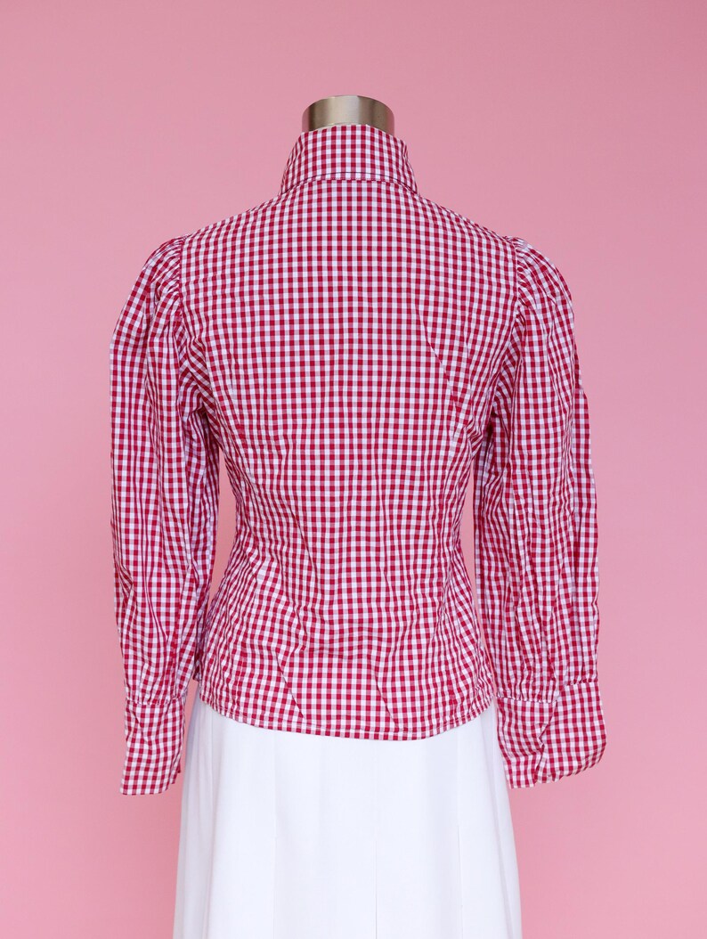 Puede incluir: Una camisa de botones de cuadros rojos y blancos con cuello alto y mangas largas. La camisa es entallada y tiene una manga ligeramente abullonada.