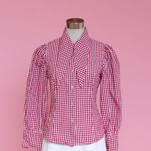 Puede incluir: Una camisa de botones de cuadros rojos y blancos con un cuello grande y puntiagudo. La camisa tiene mangas largas con hombros abullonados y una silueta ajustada.