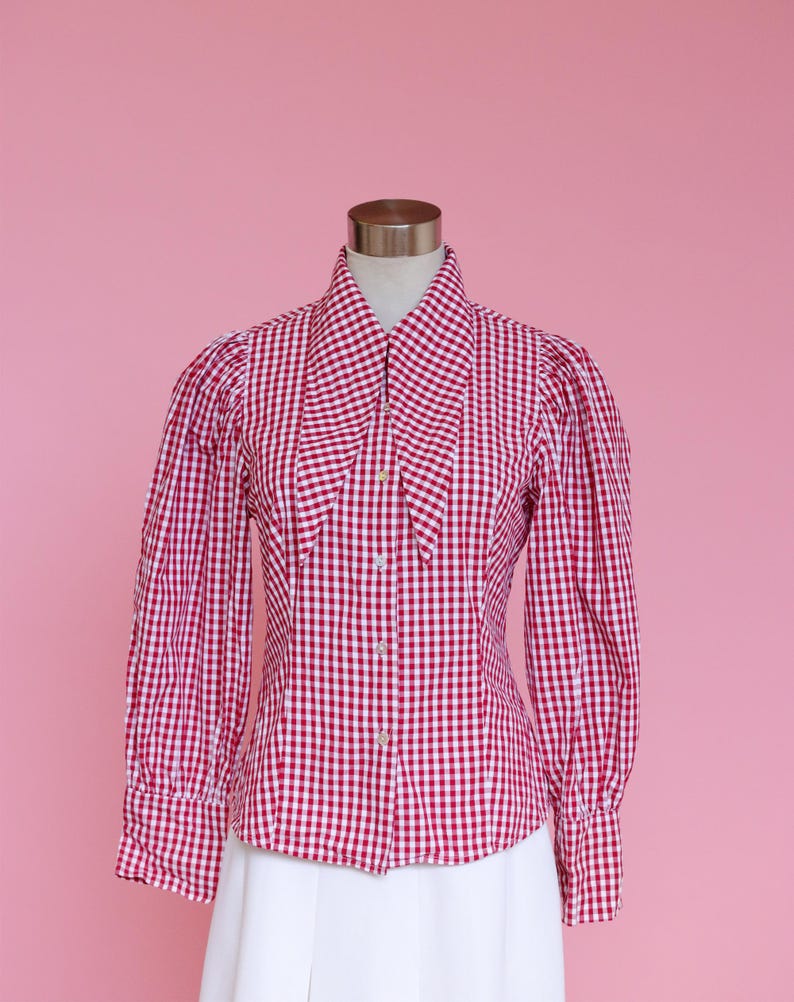 Puede incluir: Una camisa de botones de cuadros rojos y blancos con un cuello grande y puntiagudo. La camisa tiene mangas largas con hombros abullonados y una silueta ajustada.