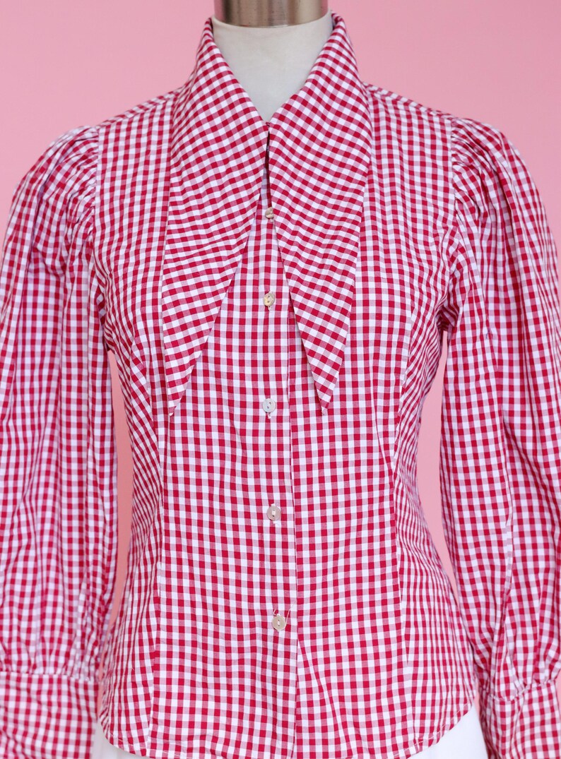 Puede incluir: Una camisa de botones de cuadros rojos y blancos con un cuello grande y puntiagudo. La camisa tiene mangas largas y una silueta ajustada.