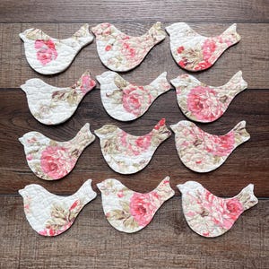 May include: Twelve quilted fabric bird cutouts arranged in four rows of three. Each bird features a white background with a repeating floral pattern in shades of pink, green, and beige. The fabric has a visible stitched texture throughout the surface.