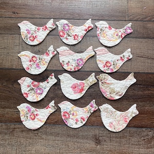 May include: Twelve quilted fabric bird-shaped cutouts arranged in four rows of three. Each piece features a white background with various floral patterns in shades of pink, red, yellow, and green. The fabric has a visible stitched texture.