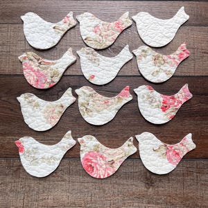 May include: A set of twelve quilted fabric bird cutouts arranged in four rows of three. Each bird shape features a white background with varying floral patterns in shades of pink, green, and beige, showcasing a textured, stitched surface.