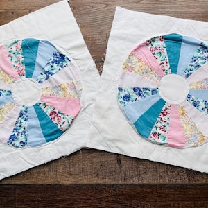 May include: Two handmade quilt blocks featuring a colorful wheel design. Each wheel is composed of fabric wedges in shades of pink, blue, teal, and floral prints, set against a white background. The blocks are placed on a wooden surface.