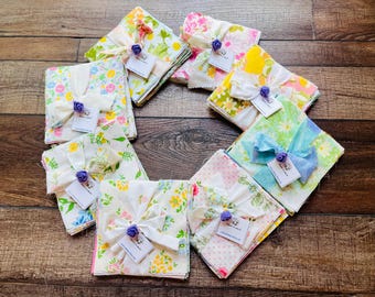 May include: A collection of eight folded fabric squares featuring various floral patterns in shades of yellow, pink, green, and blue. Each stack is tied with a white ribbon, a small purple fabric flower, and a white rectangular paper tag.