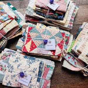 May include: Four stacks of assorted fabric quilt scraps tied with white string and decorated with a small purple fabric flower and a white tag that reads Thanks so much. The fabric pieces feature various patterns including floral, plaid, and geometric prints.