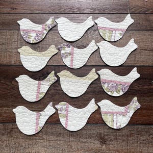 May include: A set of twelve bird-shaped fabric cutouts arranged in four rows. Each cutout features quilted white fabric with accents of pink, green, and purple floral patterns. The shapes are flat and display various combinations of these quilted textures.