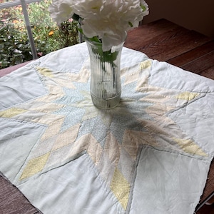 Pode incluir: Uma colcha azul clara e amarela com um padrão de estrela numa mesa de madeira. Um vaso de vidro transparente com flores brancas está no centro da colcha. A colcha tem um desenho de estrela com padrões em forma de diamante.