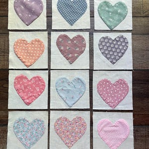 May include: Twelve square fabric patches arranged in a grid, each featuring a heart-shaped applique. The hearts display various patterns including floral, polka dot, and solid colors in shades of pink, blue, green, purple, and orange on cream fabric.