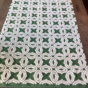 May include: A rectangular table runner featuring a solid sage green fabric base with a raised, white tufted geometric pattern. The design consists of repeating interconnected oval and diamond shapes that create a textured, soft surface.