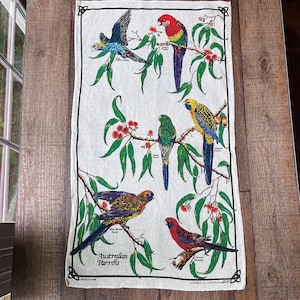 May include: A rectangular off-white linen and cotton tea towel featuring colorful illustrations of Australian parrots perched on eucalyptus branches with red flowers. The text reads Australian Parrots, Pale-headed Rosella, Eastern Rosella, Red-rumped Parrot, Adelaide Rosella, Blue-bonnet Parrot, and Crimson Rosella.