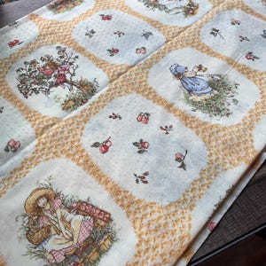 May include: A fabric featuring a repeating grid pattern of yellow wheat-like borders. The panels alternate between illustrations of children in straw hats and bonnets picking fruit and smaller squares with scattered red apple motifs.
