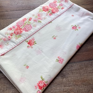 May include: A white fabric sheet featuring a prominent border of pink and red rose floral patterns. The main body of the fabric is decorated with smaller, scattered pink rose motifs. A delicate pink trim separates the border from the center.