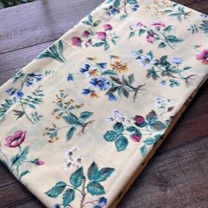 May include: A folded piece of light yellow fabric featuring a repeating botanical print of various wildflowers, green leaves, small red strawberries, and dark blue berries scattered across the surface in a detailed, colorful pattern.