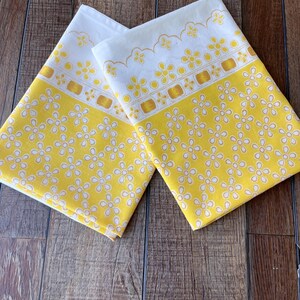 May include: Two rectangular fabric pieces featuring a bright yellow background with a repeating white five-petal flower pattern. The top section of each piece has a white border with a yellow scalloped edge and a row of yellow floral motifs.