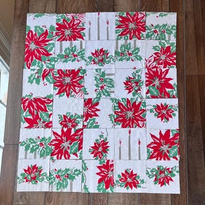 May include: A patchwork quilt featuring a repeating pattern of red poinsettias, green holly, and gray candles on a white background. The quilt is made of individual squares arranged in a grid pattern, perfect for holiday decor.