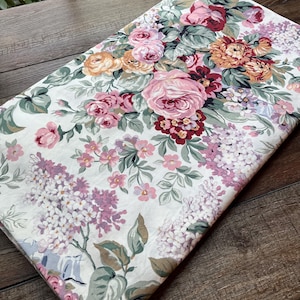May include: A folded piece of fabric featuring a floral pattern with pink, peach, and deep red roses, along with clusters of light purple lilac flowers and green leaves on a white background. The textile displays a dense, multi-colored botanical print.