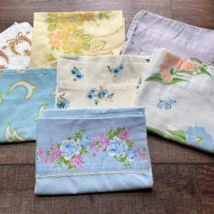May include: A collection of vintage pillowcases and sheets in various floral patterns. The colors include light blue, yellow, white, and lavender. Some feature lace trim. The sheets are folded and arranged on a dark wood surface.