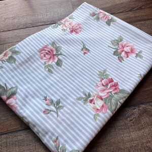 May include: A folded fabric piece featuring a light blue and white vertical striped pattern overlaid with a floral print of pink roses and green leaves. The textile has a smooth texture and is arranged in a rectangular shape on a surface.