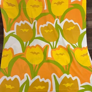 May include: A fabric textile featuring a repeating pattern of yellow and orange tulips with green stems and leaves set against a white background. The floral design is composed of bold, flat shapes with distinct outlines in a vertical layout.