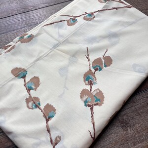 May include: A folded off-white fabric featuring a repeating pattern of brown branches with soft, rounded buds. Each bud is detailed with a central accent of bright turquoise blue. The textile has a smooth, matte finish and is neatly folded.