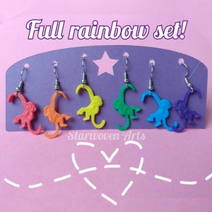 May include: A set of six monkey earrings in rainbow colors, displayed on a purple stand. The earrings are made of plastic and have a 3D monkey design. The earrings are arranged in a rainbow order, from red to purple.