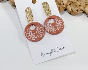 May include: A pair of dangle earrings on a white display card labeled Connaught & Carole. Each earring features a gold glitter rectangular top attached to a circular terracotta-colored clay pendant with a white hand-painted floral pattern.
