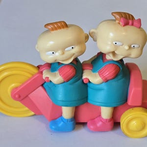 May include: A plastic toy featuring two cartoon characters with pale skin, one with a small brown tuft of hair and the other with a pink bow, riding a pink and yellow tricycle. Both characters wear blue overalls over pink shirts and blue or pink shoes.