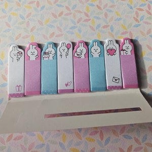 Può includere: Un set di otto post-it a tema coniglietto, ognuno con un'illustrazione diversa. I bigliettini hanno sfondi rosa e blu e sono tenuti in un supporto bianco. Perfetti per annotare note e promemoria, sono un affascinante articolo di cancelleria.