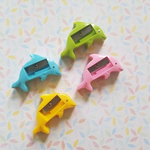 Könnte beinhalten: Vier bunt gefärbte Anspitzer in Form von Delfinen in Blau, Grün, Rosa und Gelb. Jeder Anspitzer hat eine silberne Klinge und ein lächelndes Delfin-Design. Die Anspitzer sind auf einem gemusterten Hintergrund angeordnet.