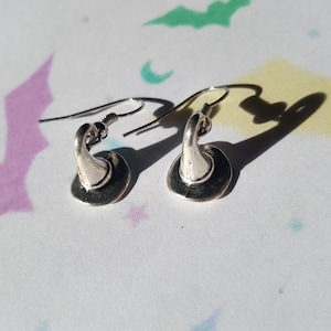 May include: A pair of silver-toned metal earrings shaped like pointed hats with wide brims. Each earring features a curved hook wire for attachment, with a polished finish that reflects light, creating distinct shadows on the surface below.