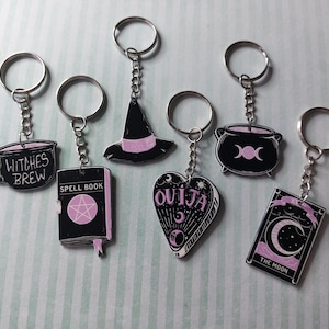 May include: A set of six black and light pink keychains featuring occult-themed designs including a mug labeled Witches Brew, a Spell Book, a witch hat, a heart-shaped Ouija planchette, a cauldron with a triple moon symbol, and a Moon tarot card.