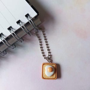 Può includere: Un ciondolo originale con un design a uovo fritto su pane tostato. Il ciondolo è quadrato, con un bordo di pane tostato marrone e un uovo bianco e arancione. È appeso a una catena a sfera argentata, vicino a un taccuino a spirale.