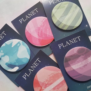 Può includere: Cinque note adesive colorate a forma di pianeta con la parola "PLANET" stampata su ciascuna. I pianeti sono rosa, verde, blu, viola e arancione con strisce bianche.