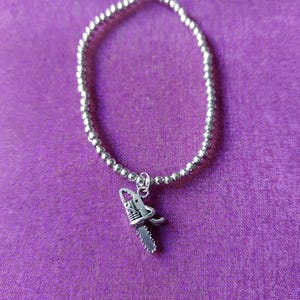 Op de afbeelding: Een armband van kleine, zilverkleurige metalen kralen met een gedetailleerde metalen bedel in de vorm van een kettingzaag. De armband bestaat uit een doorlopende lus van uniforme metalen bolletjes, waaraan de kettingzaagbedel is bevestigd.