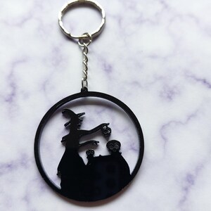 May include: A black circular keychain featuring a silhouette of a witch standing beside a cauldron with two skulls. The design is attached to a silver-colored metal split ring by a short chain, creating a cohesive accessory with a dark aesthetic.