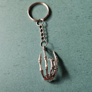 May include: A silver-toned metal keychain featuring a skeletal hand charm. The charm is shaped like a hand with fingers curled inward, attached to a standard circular split ring by a short metal chain. The finish is polished and metallic.