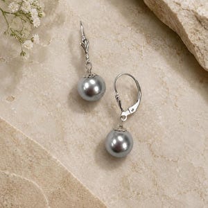 May include: A pair of silver leverback earrings with large, round, gray pearl drops. The earrings are displayed on a light beige surface, with a sprig of white flowers in the upper left corner.
