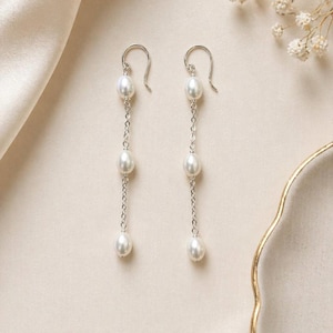 May include: Elegant dangle earrings featuring three lustrous white pearls suspended from delicate silver chains. The earrings have a classic design with a hook closure, perfect for adding a touch of sophistication to any outfit.
