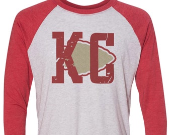 Puede incluir: Una camiseta de estilo béisbol roja y blanca con un logotipo "KC" rojo desgastado con una punta de flecha beige dentro de la "C".