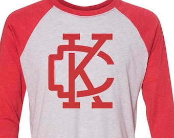 May include: A red and white baseball style t-shirt with a red "KC" logo on the front.