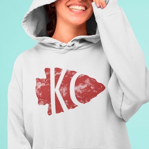 May include: White hoodie with a red distressed arrowhead design featuring the letters "KC" in white.