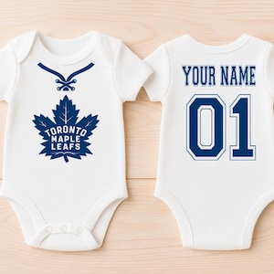 Puede incluir: Body de bebé blanco con el logotipo azul de los Toronto Maple Leafs y el texto "TORONTO MAPLE LEAFS". La parte trasera del body tiene el texto "YOUR NAME" encima del número "01" en azul.