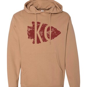 May include: A tan hoodie with a red distressed arrowhead graphic featuring the letters "KC" inside.