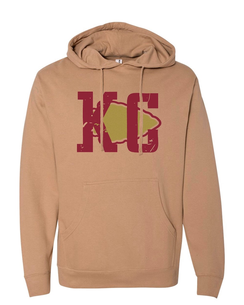 May include: A tan hoodie with a red and gold graphic of the letters "KC" with an arrowhead in the middle.
