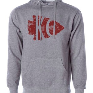 May include: A gray hooded sweatshirt with a red distressed arrowhead design featuring the letters "KC" inside.