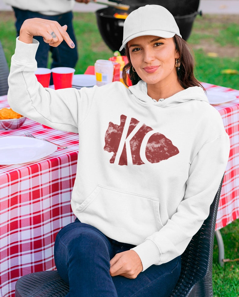 May include: White hoodie with a red distressed graphic of the letters "KC" inside a state outline. The model is wearing a white baseball cap and blue jeans.