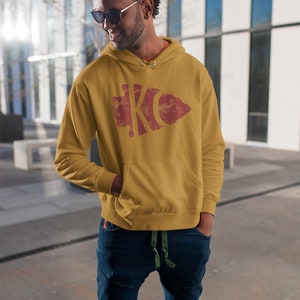 May include: A mustard yellow hoodie with a red and white distressed graphic of the letters "KC" inside an arrowhead shape.