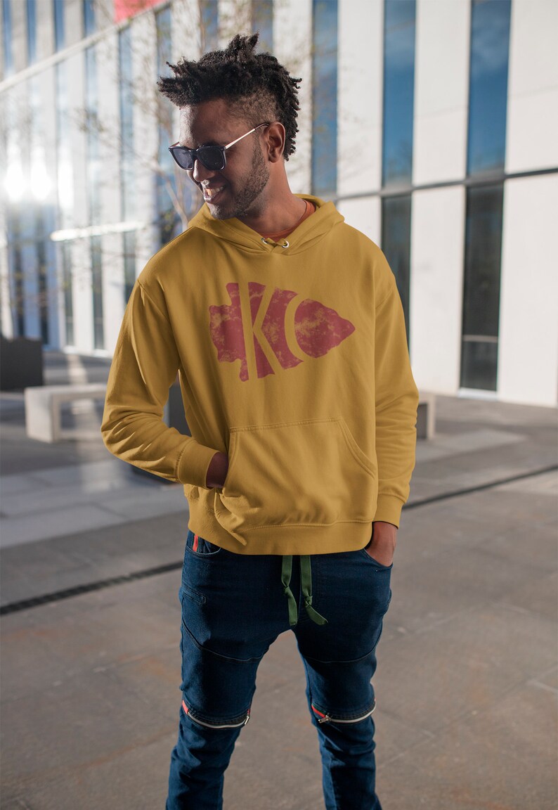 May include: A mustard yellow hoodie with a red and white distressed graphic of the letters "KC" inside an arrowhead shape.