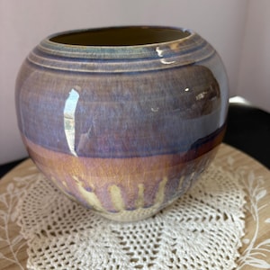 May include: A round ceramic vase with a wide opening. The vase has a glossy finish with colors that transition from purple to gold. The vase sits on a white lace doily and a wooden tray.